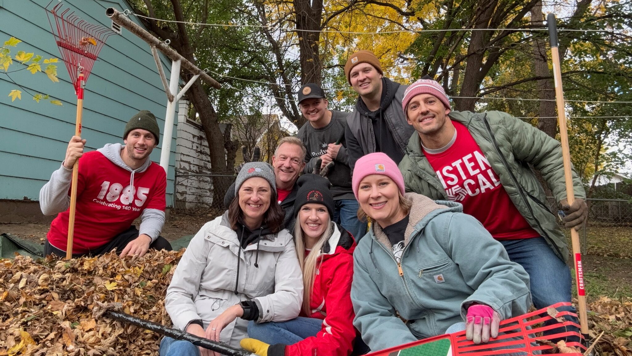 A group of smiling volunteers pose with rakes and a pile of leaves