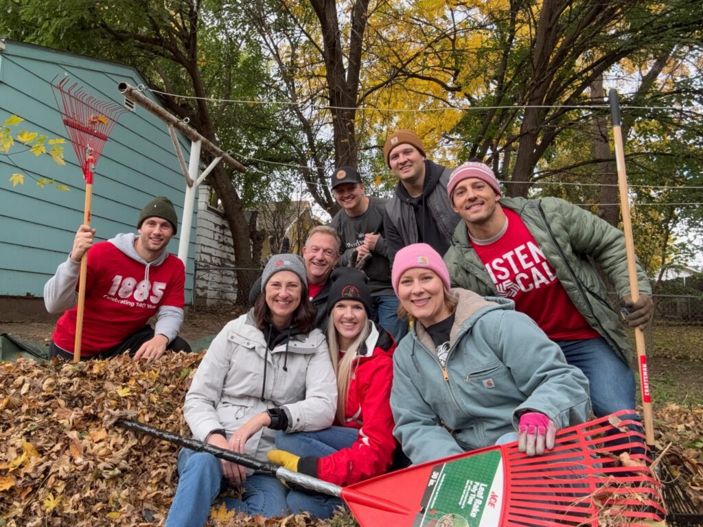 A group of smiling volunteers pose with rakes and a pile of leaves